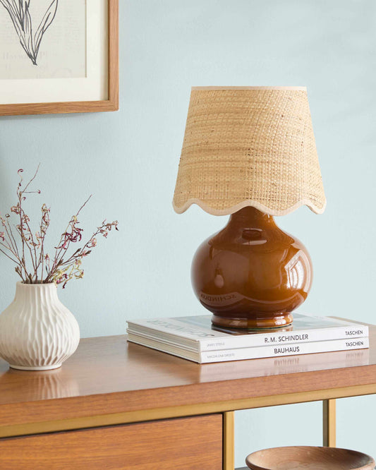 A table lamp with a glossy amber-brown ceramic base and a natural woven beige lampshade, styled on a stack of books on a wooden console table against a light blue wall.