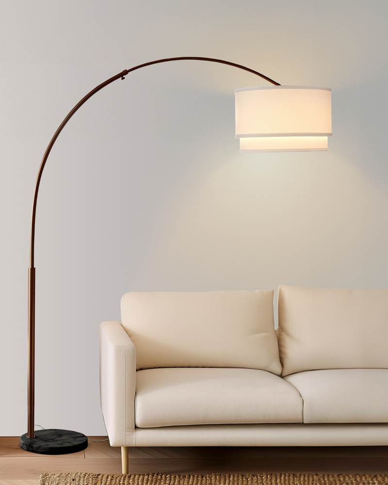 An arched floor lamp with a dark bronze metal frame and a white drum shade, shown lit next to a cream-colored sofa in a minimalist living room setting.