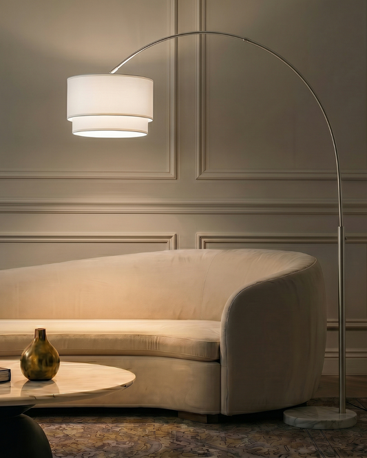 An Arc Light floor lamp with a curved silver metal stand and a white drum shade, glowing warmly in a modern living room setting next to a beige sofa.